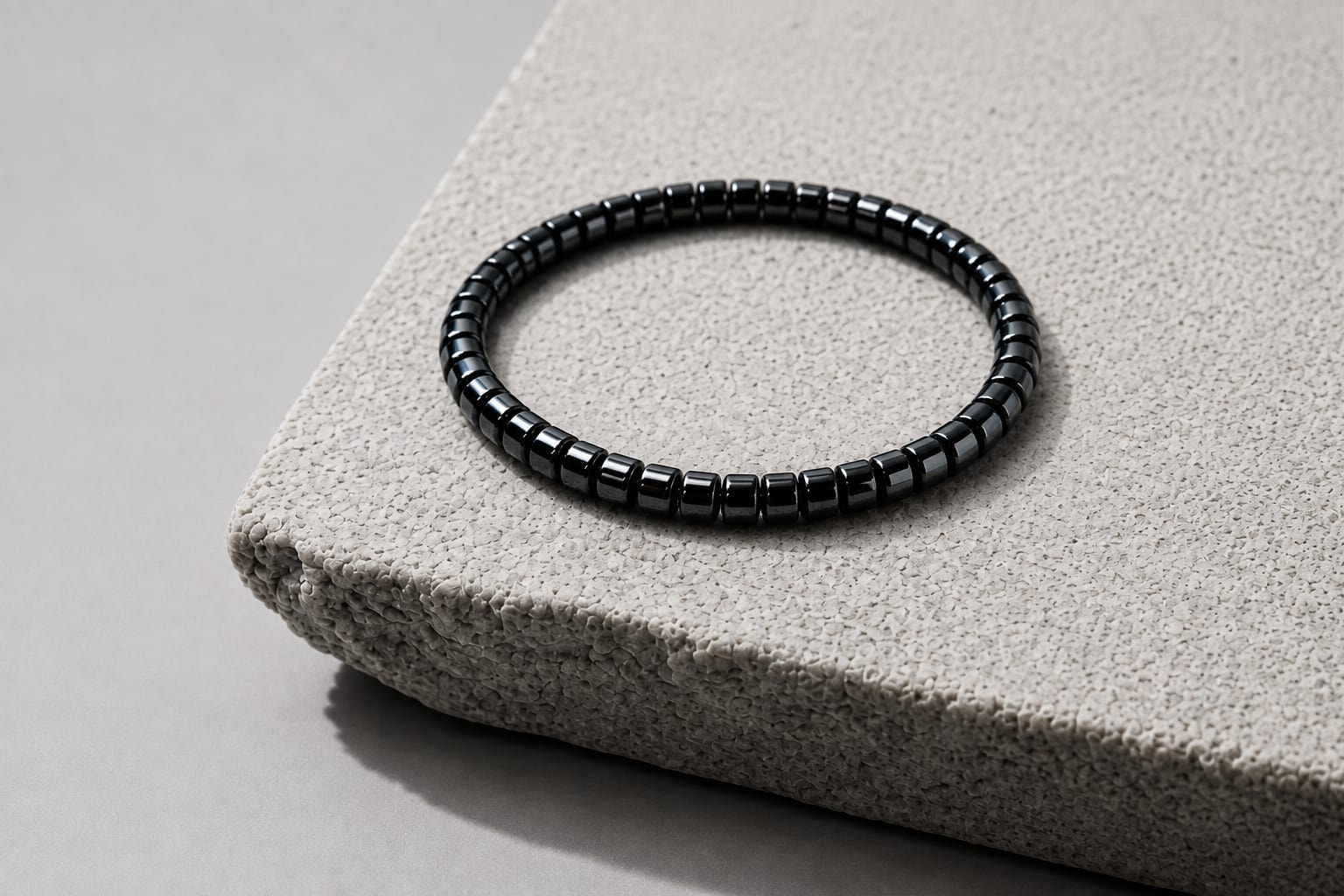 The IONISS hematite bracelet resting on the corner of a pale textured stone plinth, its polished gunmetal beads lit softly against a grey background