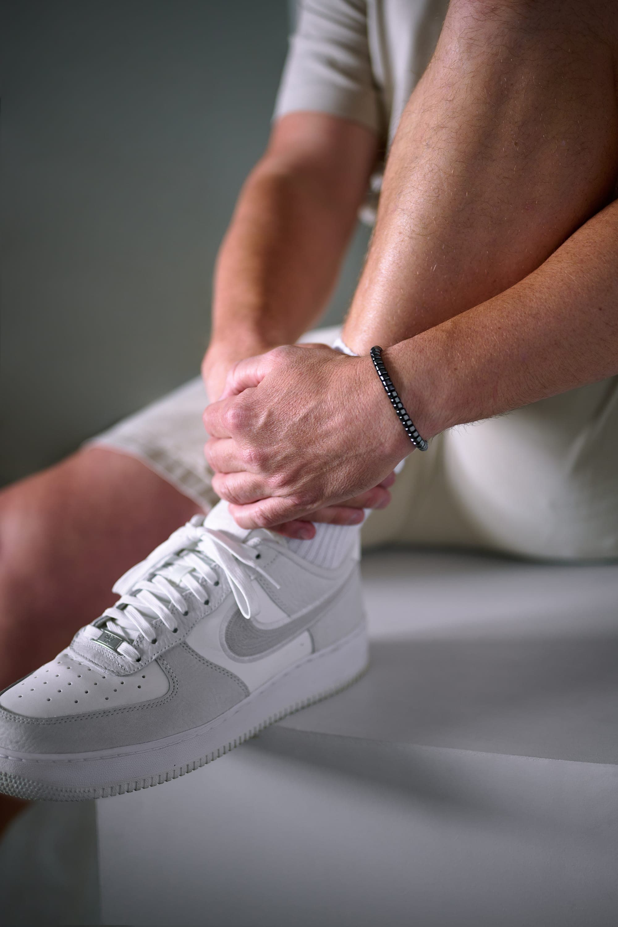 A man seated in shorts and white trainers, the IONISS bracelet on his wrist as he ties a lace